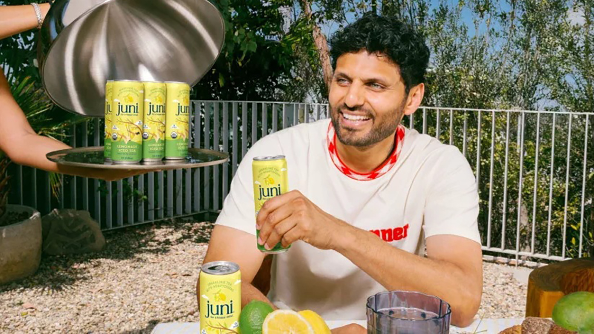 A man holds a Juni Lemonade Tea can and smiles while more cans are served on a tray outdoors.